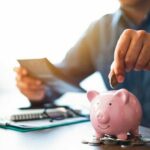 Close-up_image_of_man_hand_putting_coins_in_pink_piggy_bank_for_account_save_money._Planning_step_up,_saving_money_for_future_plan,_retirement_fund._Business_investment-finance_accounting_concept.