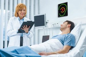Friendly_Female_Doctor_Visits_Happy_Recovering_Man_who_is_Lying_in_Bed,_She_Asks_Him_Questions_and_Fills_Medical_Chart._Friendly_Doctor_and_Sick_Man_in_a_Clean_Hospital_Ward.
