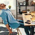 Side_view_of_freelance_male_graphic_designer_sitting_at_table_with_large_monitor_and_gadgets_and_relaxing_after_working_hard_on_project_