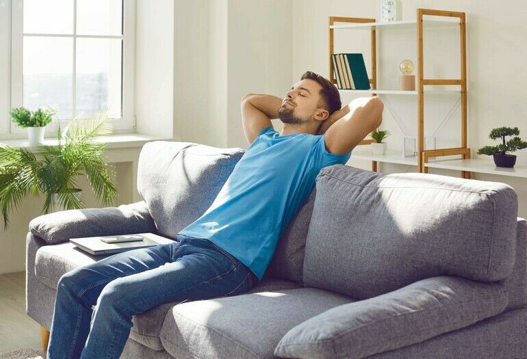 Man_relaxing_on_a_comfortable_couch_at_home._Young_guy_in_a_blue_T_shirt_sitting_on_a_gray_couch_with_his_hands_behind_his_head_and_a_laptop_beside_him._Relaxation,_break,_pause,_free_time_concept