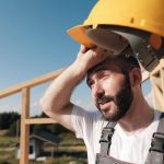 The_man_is_a_builder_on_the_background_of_the_roof_of_a_frame_house,_in_a_yellow_helmet_and_gray_overalls.
