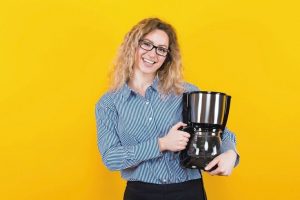 Portrait_of_attractive_curly-haired_woman_in_striped_shirt_and_eyeglasses_isolated_on_orange_background_holding_coffee_machine_advertising_concept.