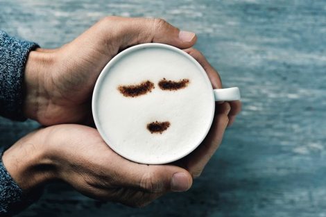 high-angle_shot_of_a_young_caucasian_man_holding_a_cup_of_cappuccino_with_a_happy_face_drawn_with_cocoa_powder_on_the_milk_foam,_on_a_gray_rustic_table