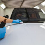 The_process_of_applying_a_nano-ceramic_coating_on_the_white_car's_hood_by_a_male_worker_in_blue_gloves_with_a_sponge_and_chemical_composition_to_protect_the_paint_in_detailing_workshop._Auto_service.