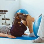 Teenage_girl_after_car_accident_with_orthosis_on_the_leg_exercises_on_blue_mat_in_the_company_of_physiotherapist