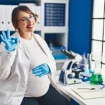 Pregnant_woman_working_at_scientist_laboratory_smiling_positive_doing_ok_sign_with_hand_and_fingers._successful_expression